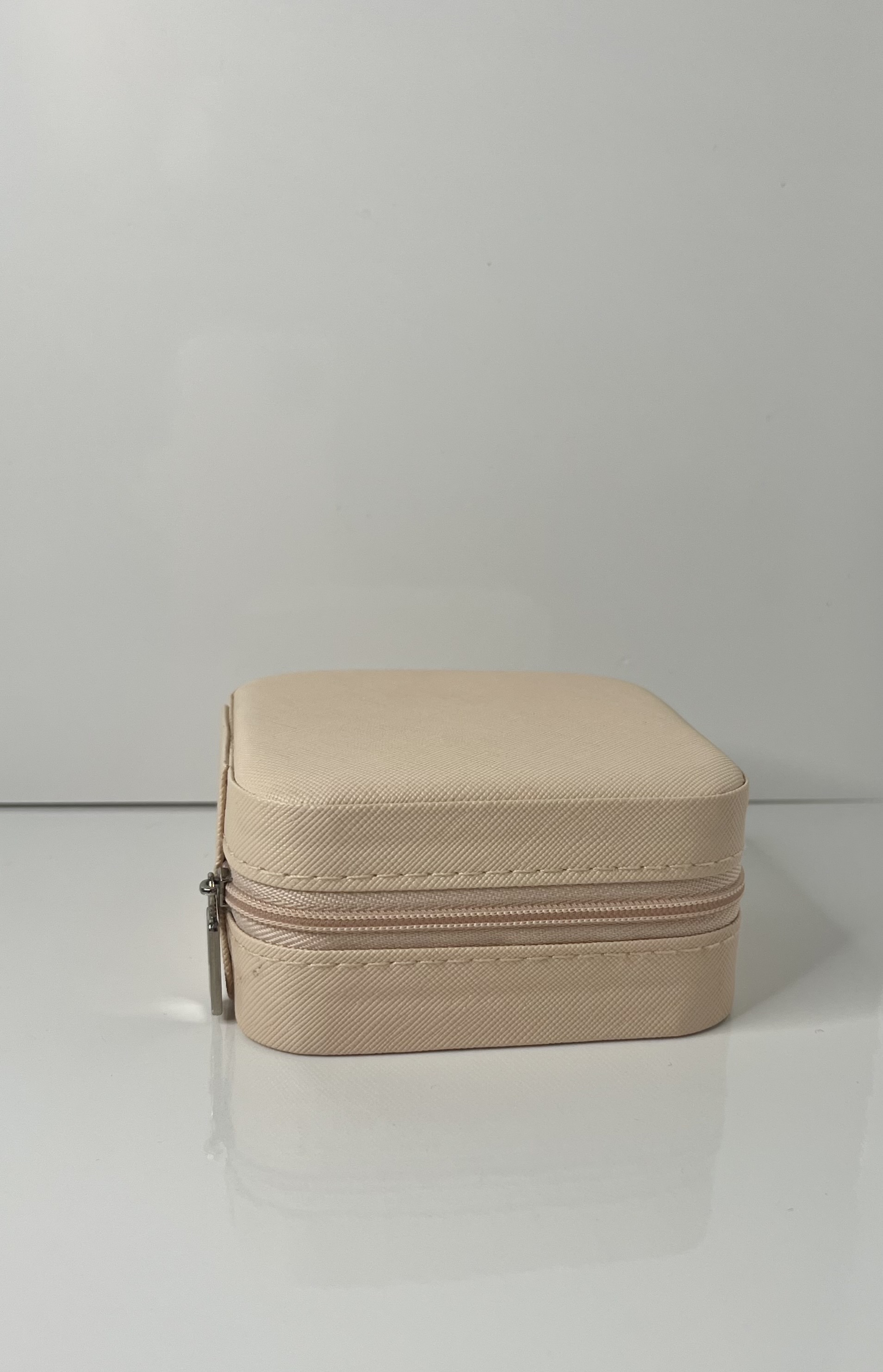 A small beige jewelry box with a zipper sits upright, its cube-like structure emphasized by clean edges and a soft fabric texture.
