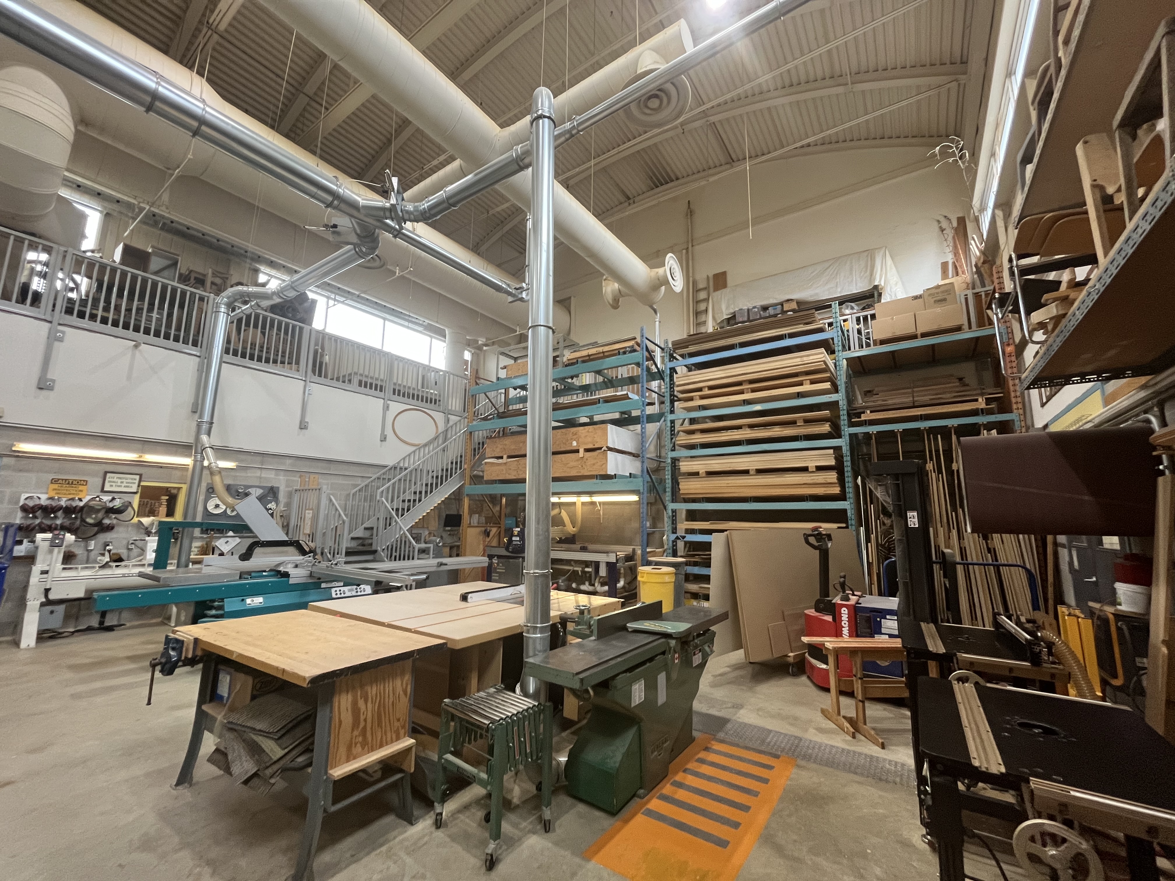 A second perspective of the woodshop reveals dust-collection systems, stacked material racks, and large-format saw equipment used in material processing.