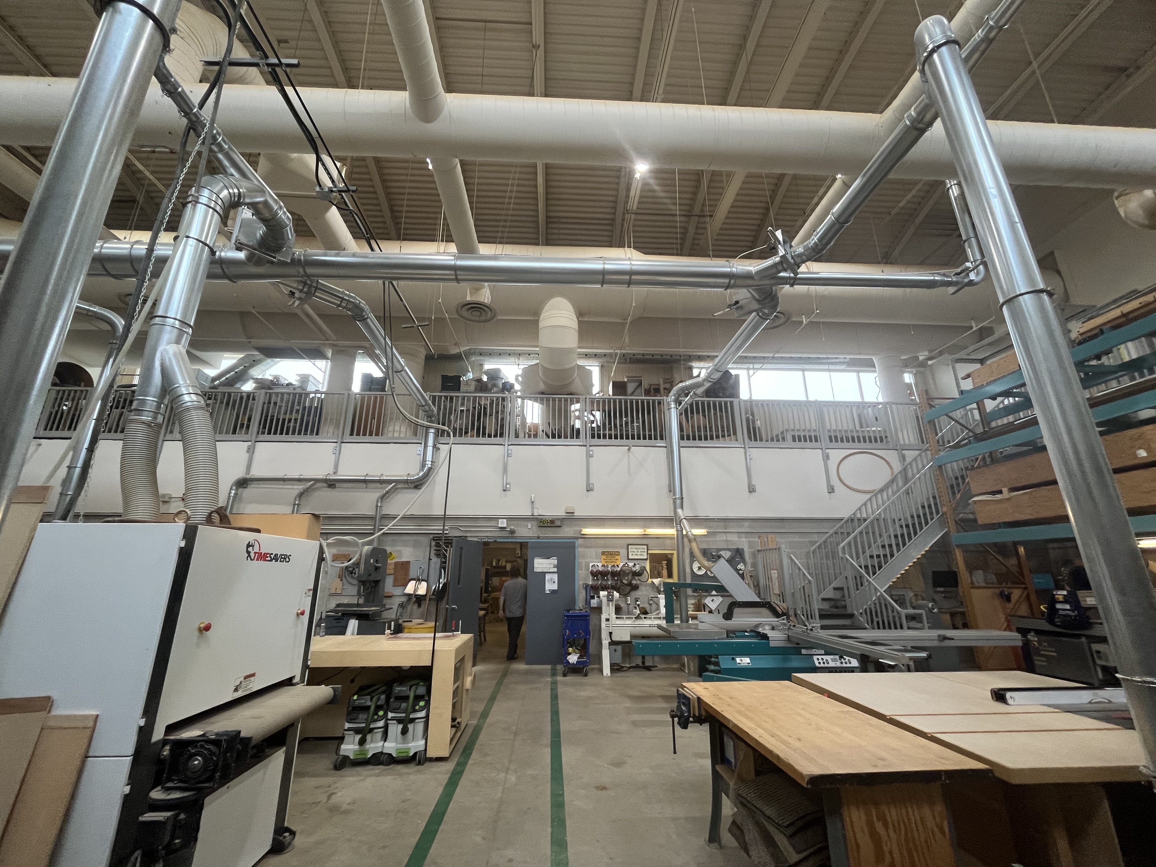The woodshop space features extensive dust-collection piping, industrial fabrication equipment, and an elevated mezzanine for additional workspace.